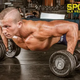 Mężczyzna w pozycji pompki, opierający się na hantlach w siłowni. Widoczne napięcie mięśni, pot na ciele i ciężkie hantle z napisem 'Haryzma Sport 32.5 kg'. W tle regał z hantlami i napis 'Sport...