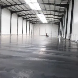 Błyszcząca, szara posadzka przemysłowa w przestronnym, pustym magazynie z białymi ścianami i stalową konstrukcją dachu, oświetlona światłem dziennym wpadającym przez świetliki dachowe.