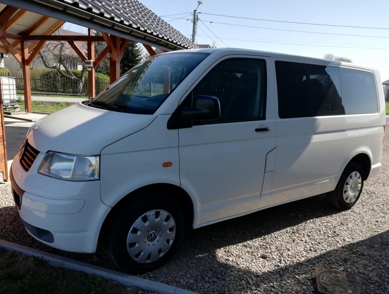Biały bus Volkswagen Transporter T5 zaparkowany na żwirowym podjeździe przed domem z drewnianą wiatą.