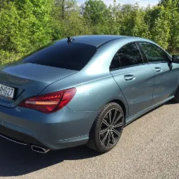 Niebieski sedan marki Mercedes-Benz CLA z widocznymi felgami aluminiowymi o wzorze wieloramiennym, zaparkowany na asfalcie z zielonym tłem drzew.