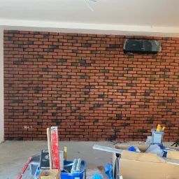 Ściana wykończona czerwoną cegłą klinkierową z czarną fugą, na ścianie zamontowany klimatyzator w folii ochronnej, widoczne narzędzia i materiały budowlane na podłodze, częściowo położona okładzina.