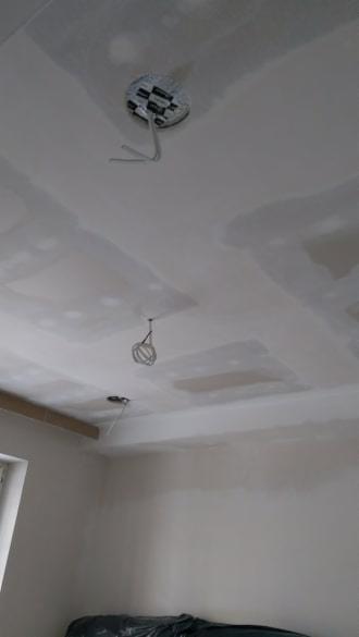 Sufit z płyt kartonowo-gipsowych w trakcie szpachlowania łączeń, widoczne przewody oświetleniowe i fragment okna z lewej strony.