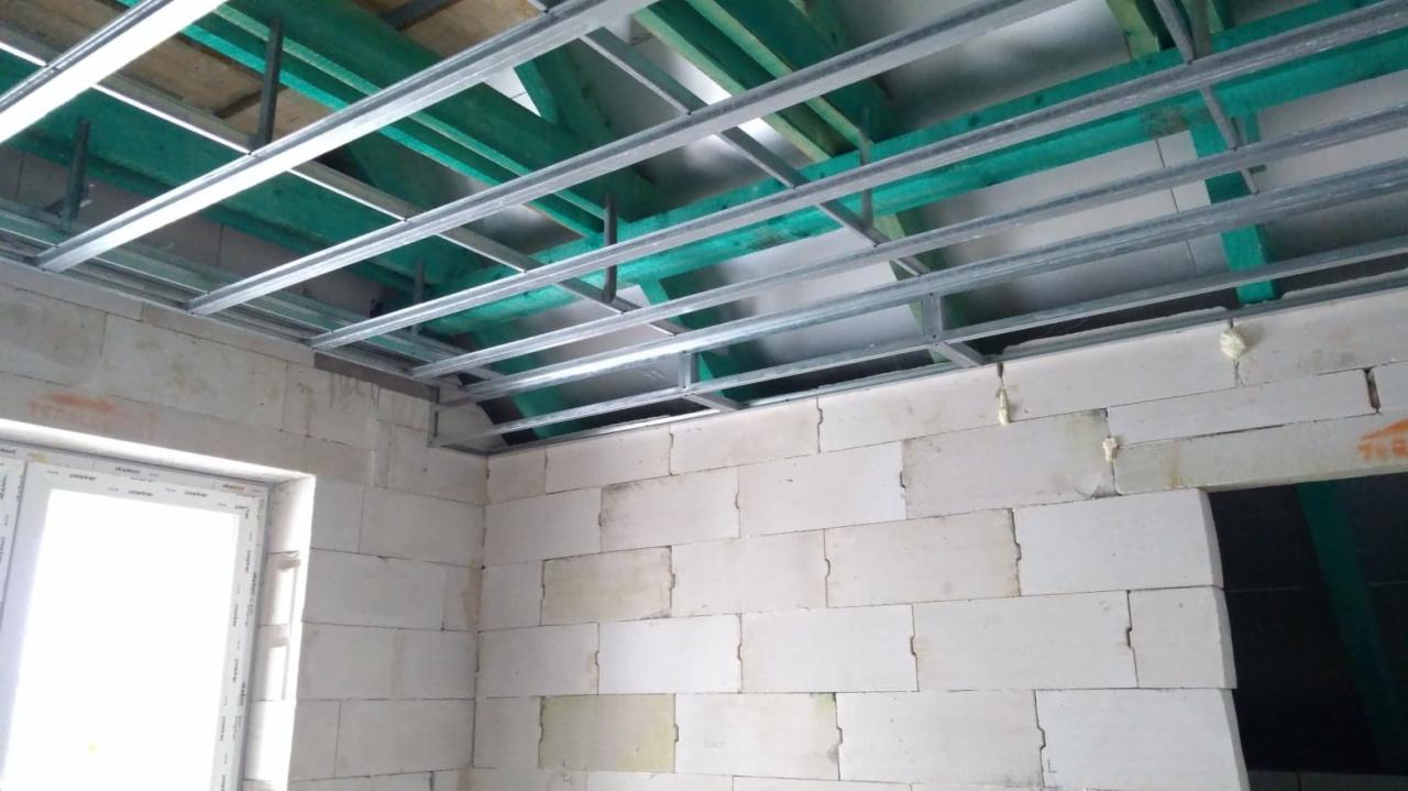 Konstrukcja metalowa podwieszanego sufitu zamontowana do drewnianego stropu, widoczne białe ściany z bloczków z betonu komórkowego i okno PCV.