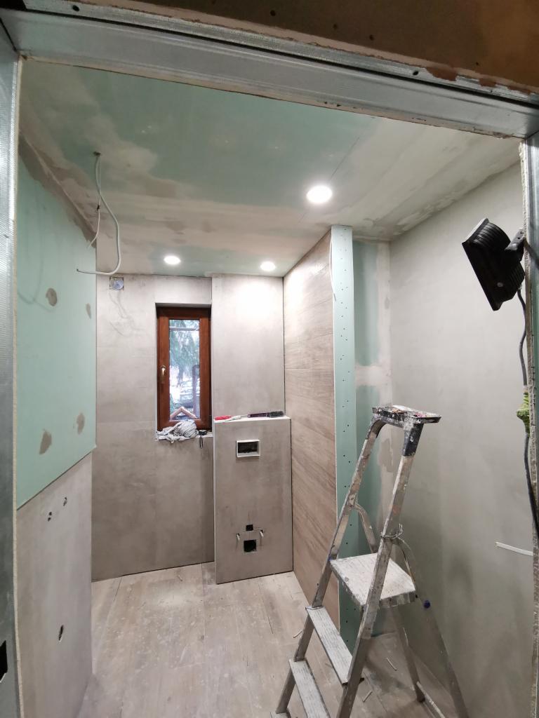 Wnętrze łazienki w trakcie remontu: widoczne płyty kartonowo-gipsowe na ścianach, szpachlowana sufit, drabina malarska, okno z drewnianą ramą i częściowo ułożona podłoga.