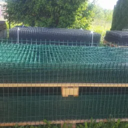 Stosy zielonych i czarnych paneli ogrodzeniowych zgrzewanych, ułożone na paletach na zewnątrz, widoczne trawa i drzewa w tle.