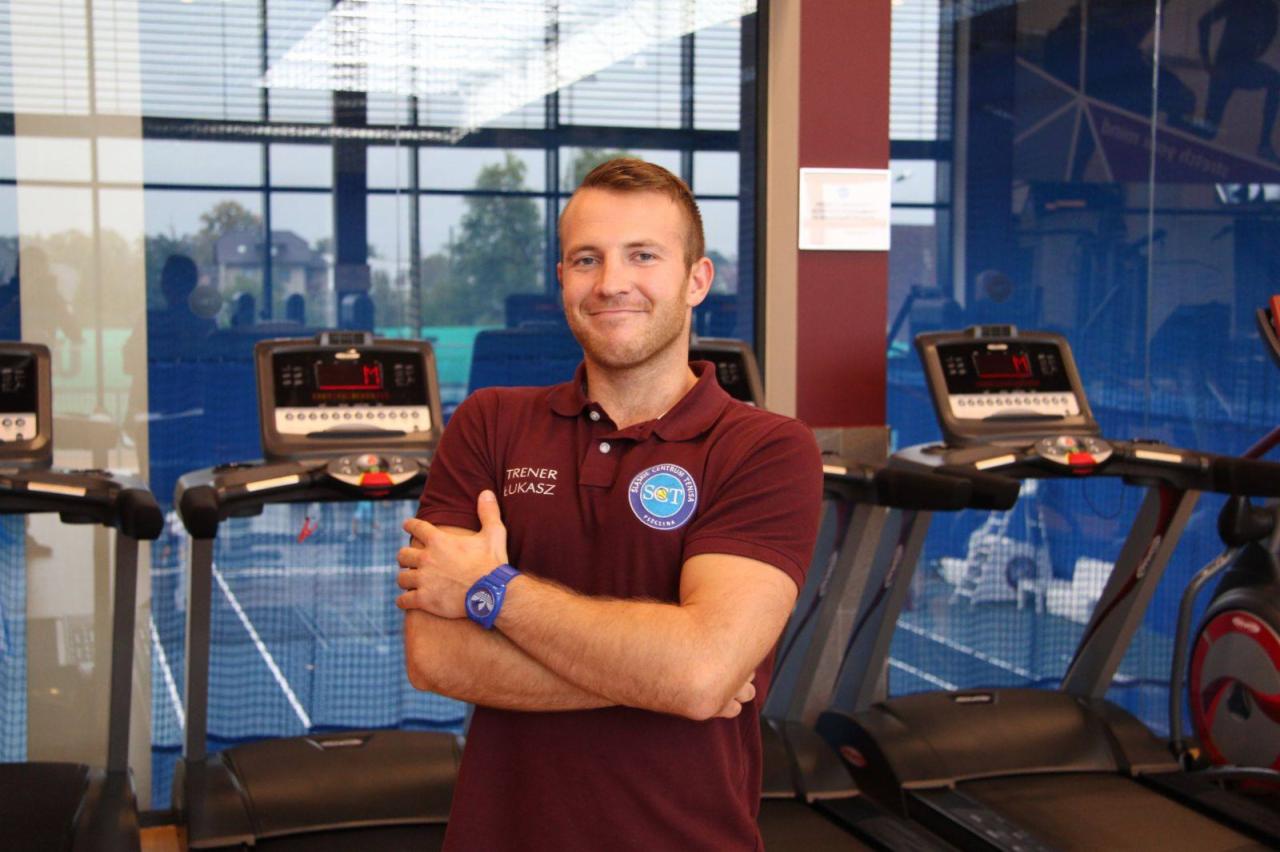 Uśmiechnięty mężczyzna w bordowej koszulce polo z napisem 'TRENER ŁUKASZ' i logo klubu sportowego, stoi na tle rzędu bieżni w siłowni, z założonymi rękami i niebieskim zegarkiem na nadgarstku.