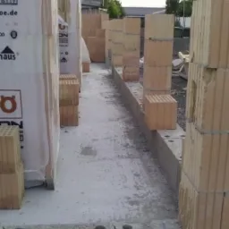 Wznoszenie ścian domu z pustaków ceramicznych Porotherm, widoczne palety z zapakowanymi bloczkami, ściany fundamentowe z otworami, przygotowane do dalszych prac.