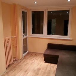 Mariusz i Adrian - Salon po remoncie: Jasne ściany, panel podłogowy, brązowa sofa narożna, białe okno i drzwi balkonowe, różowy grzejnik z rurami na ścianie.