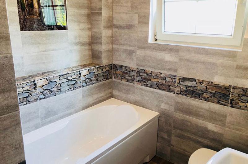Wykończona łazienka z wanną, szare płytki imitujące kamień, dekoracyjny pas z motywem kamieni łupanych, okno z białymi ramami i obrazek z motywem uliczki na ścianie.