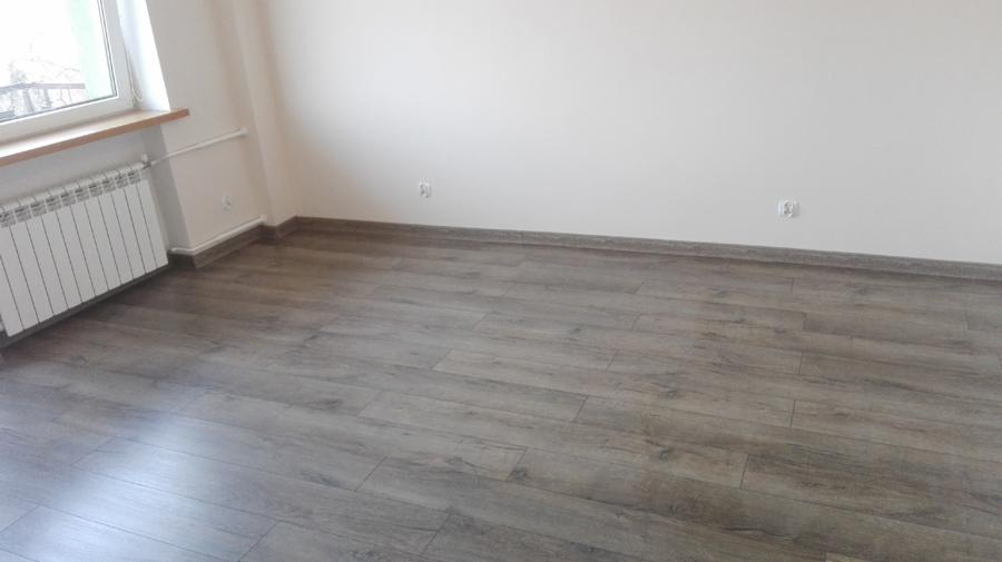 Szare panele podłogowe ułożone w pustym, odnowionym pokoju z białymi ścianami i grzejnikiem pod oknem.