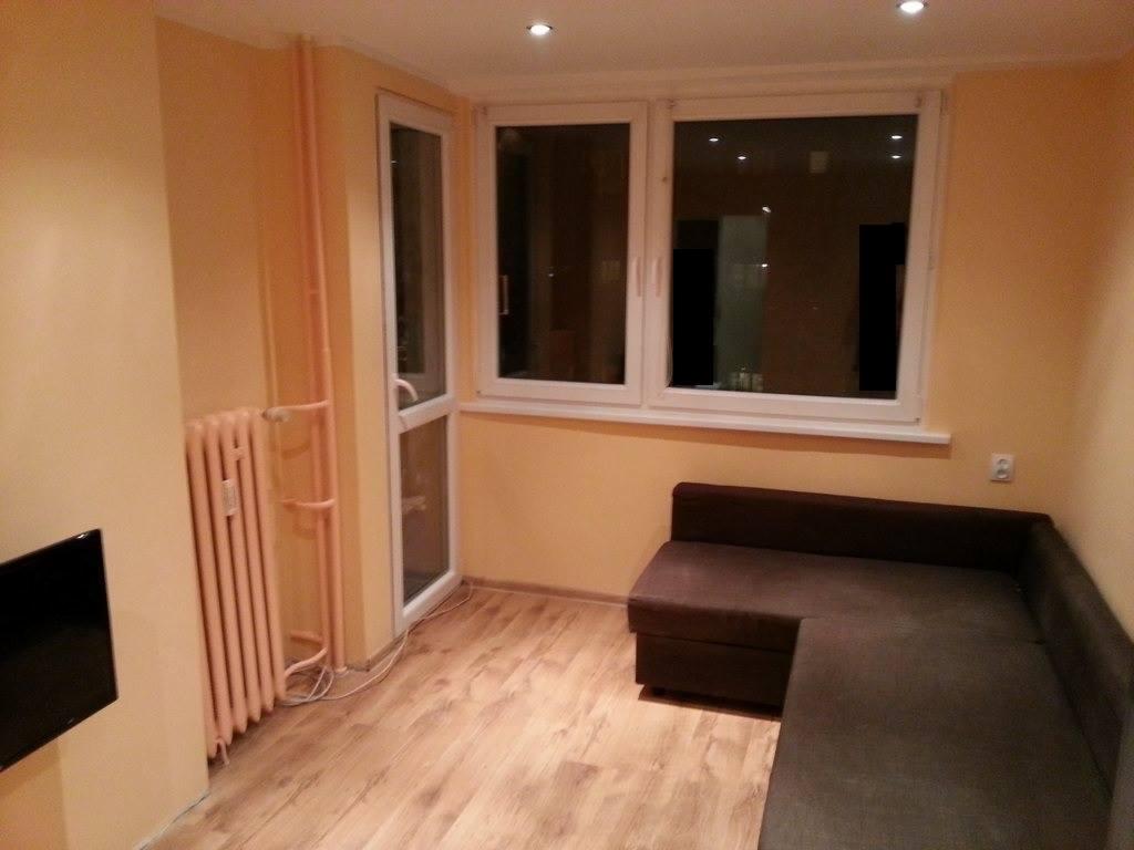 Salon po remoncie: Jasne ściany, panel podłogowy, brązowa sofa narożna, białe okno i drzwi balkonowe, różowy grzejnik z rurami na ścianie.