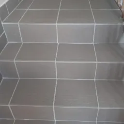 Schody wyłożone szarymi płytkami ceramicznymi z poziomymi rowkami na krawędziach stopni, wykończone jasną fugą.