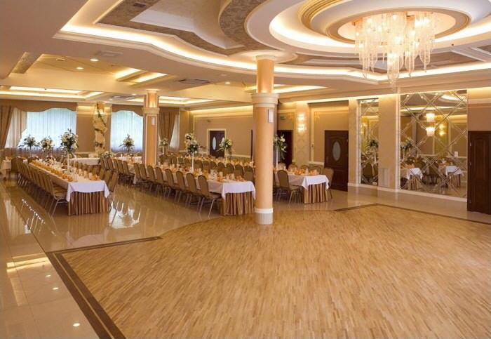 Uroczysta sala balowa z parkietem, długimi stołami nakrytymi białymi obrusami, dekoracjami kwiatowymi i kryształowym żyrandolem.