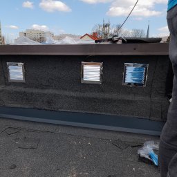 Michał - Płaski dach pokryty papą z trzema nowymi, metalowymi wywietrznikami, widok z góry, w tle budynki mieszkalne i wieża kościoła.