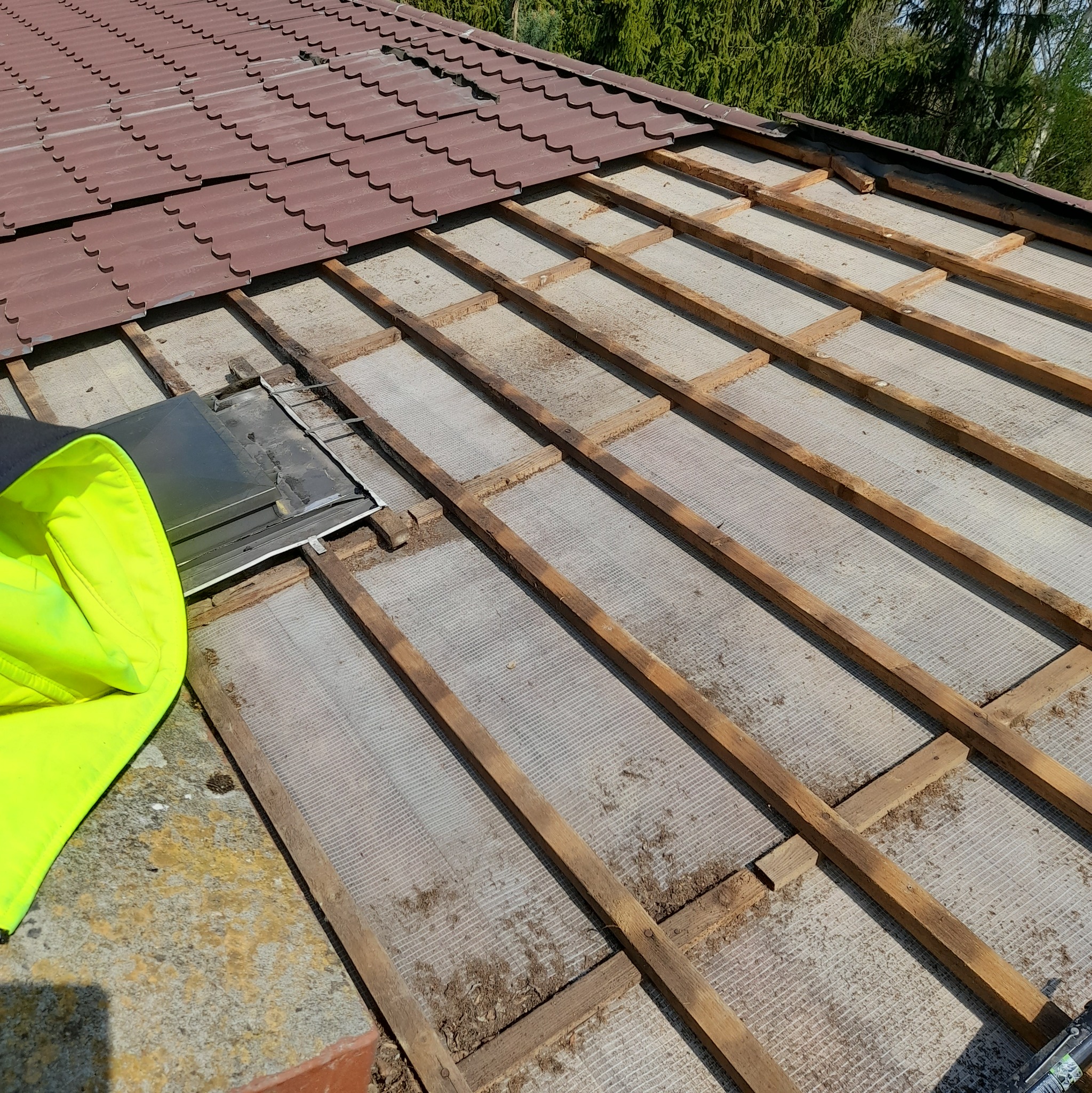Demontaż brązowej dachówki ceramicznej, widoczne drewniane łaty i folia dachowa, obok leży odblaskowa kurtka robocza