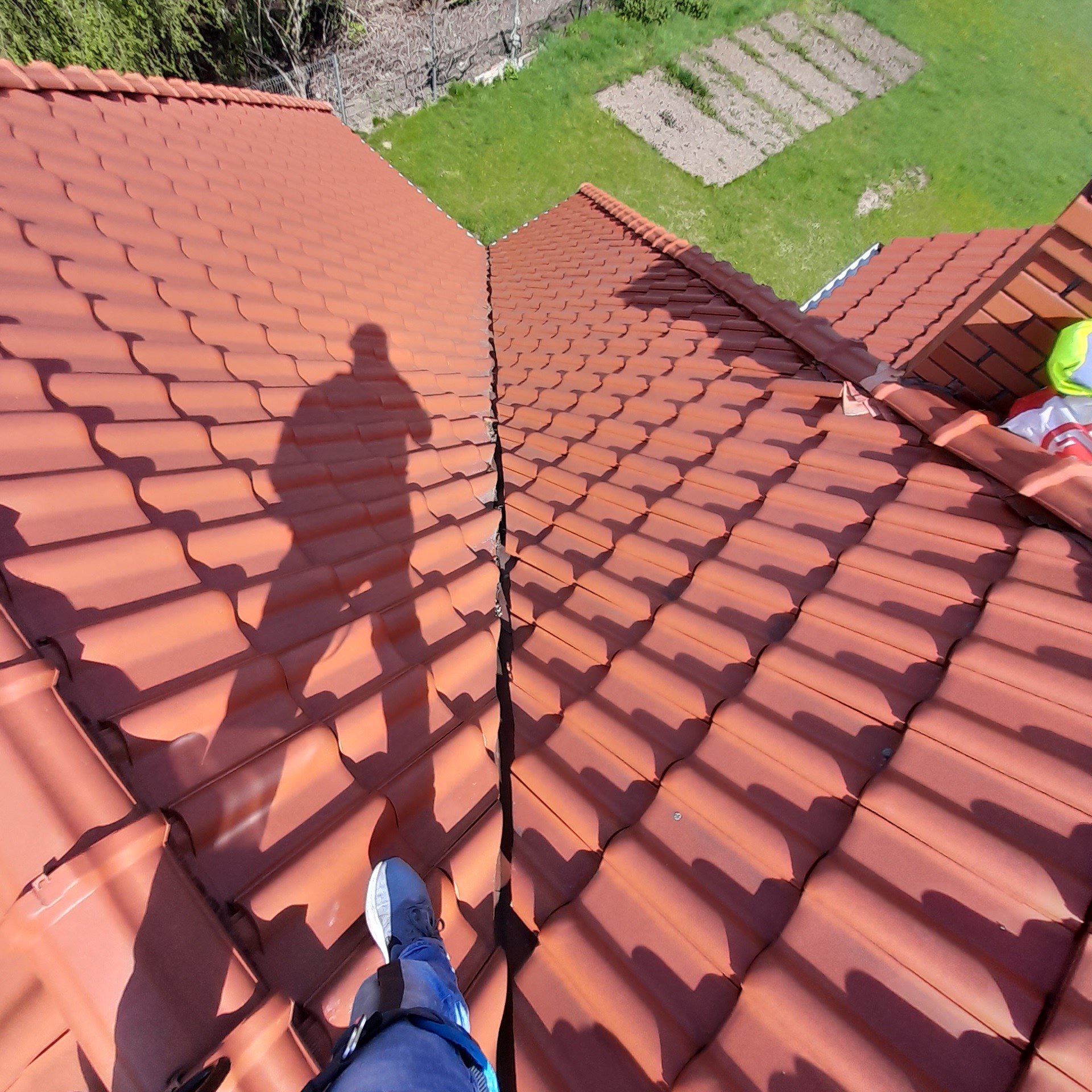 Czerwona dachówka karpiówka z widocznym pęknięciem na łączeniu połaci, cień osoby na dachu, w tle trawnik i grządki.