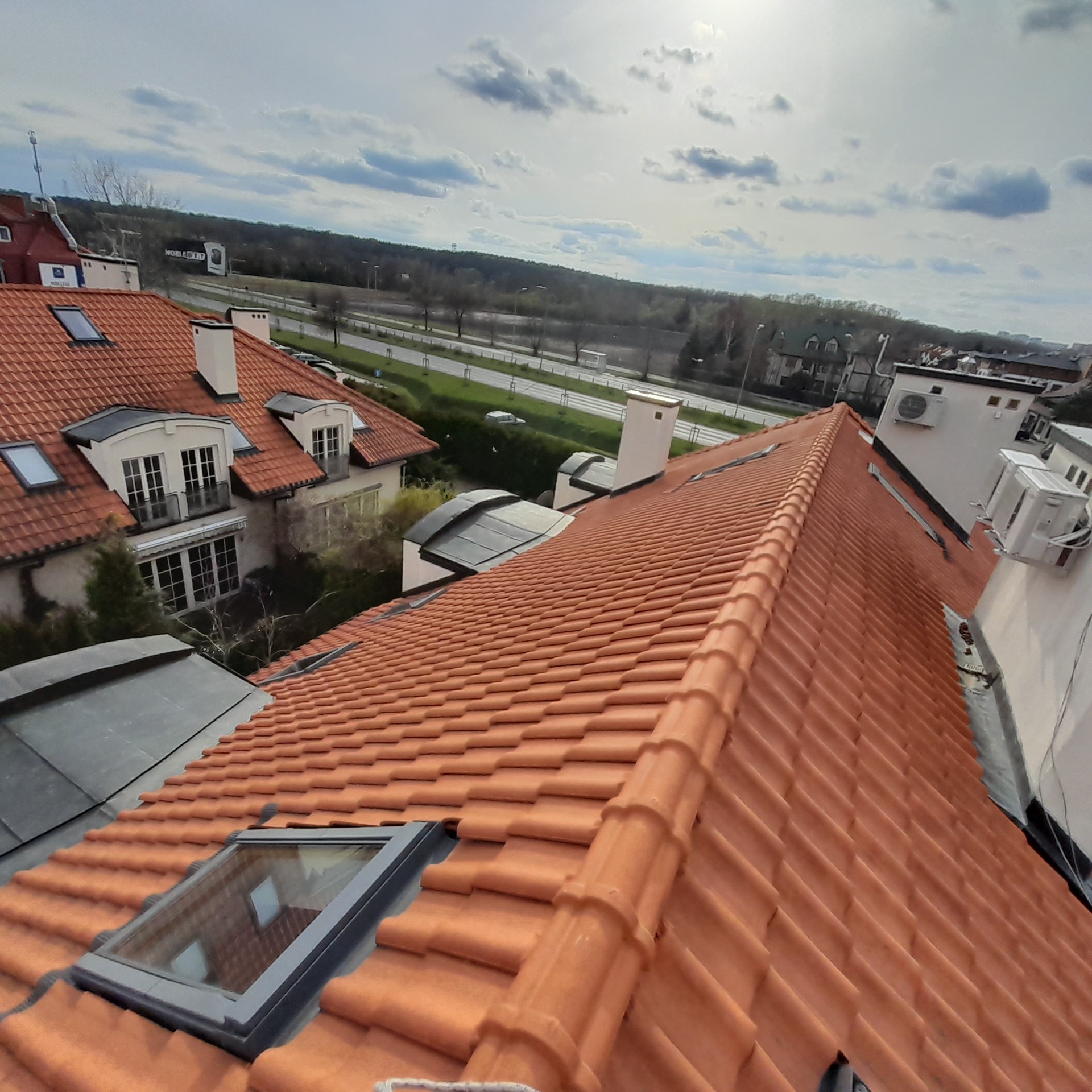 Czerwona dachówka ceramiczna na skośnym dachu z oknami dachowymi i kominami, widok z góry na budynki mieszkalne i ulicę w tle.