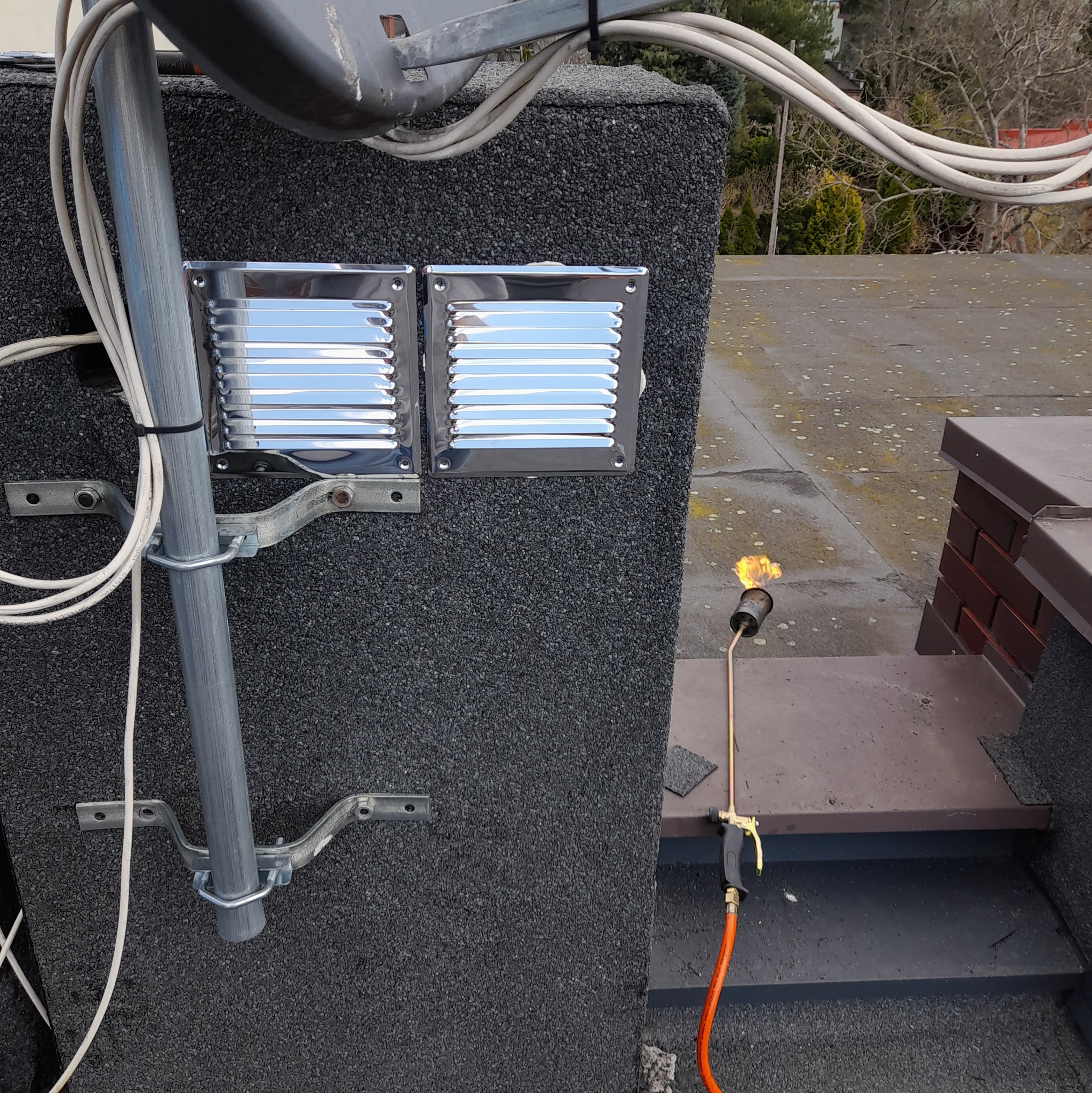 Prace dekarskie na dachu pokrytym papą: palnik dekarski w użyciu obok komina, widoczne elementy wentylacyjne i instalacja odgromowa.