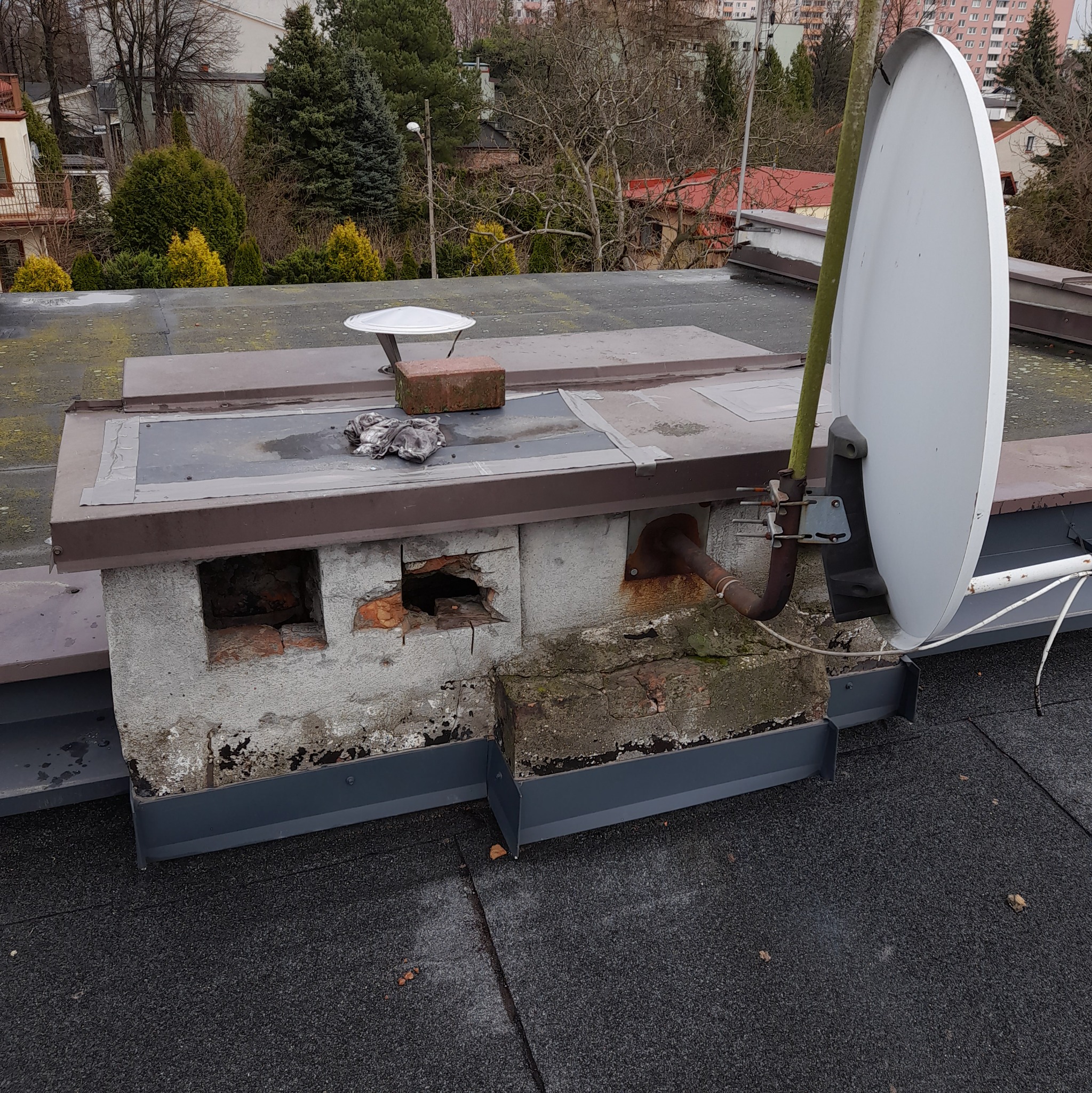 Płaski dach pokryty papą z uszkodzonym kominem z cegły i betonu, na którym zamontowana jest antena satelitarna. Widoczne ubytki w kominie i ślady wilgoci.