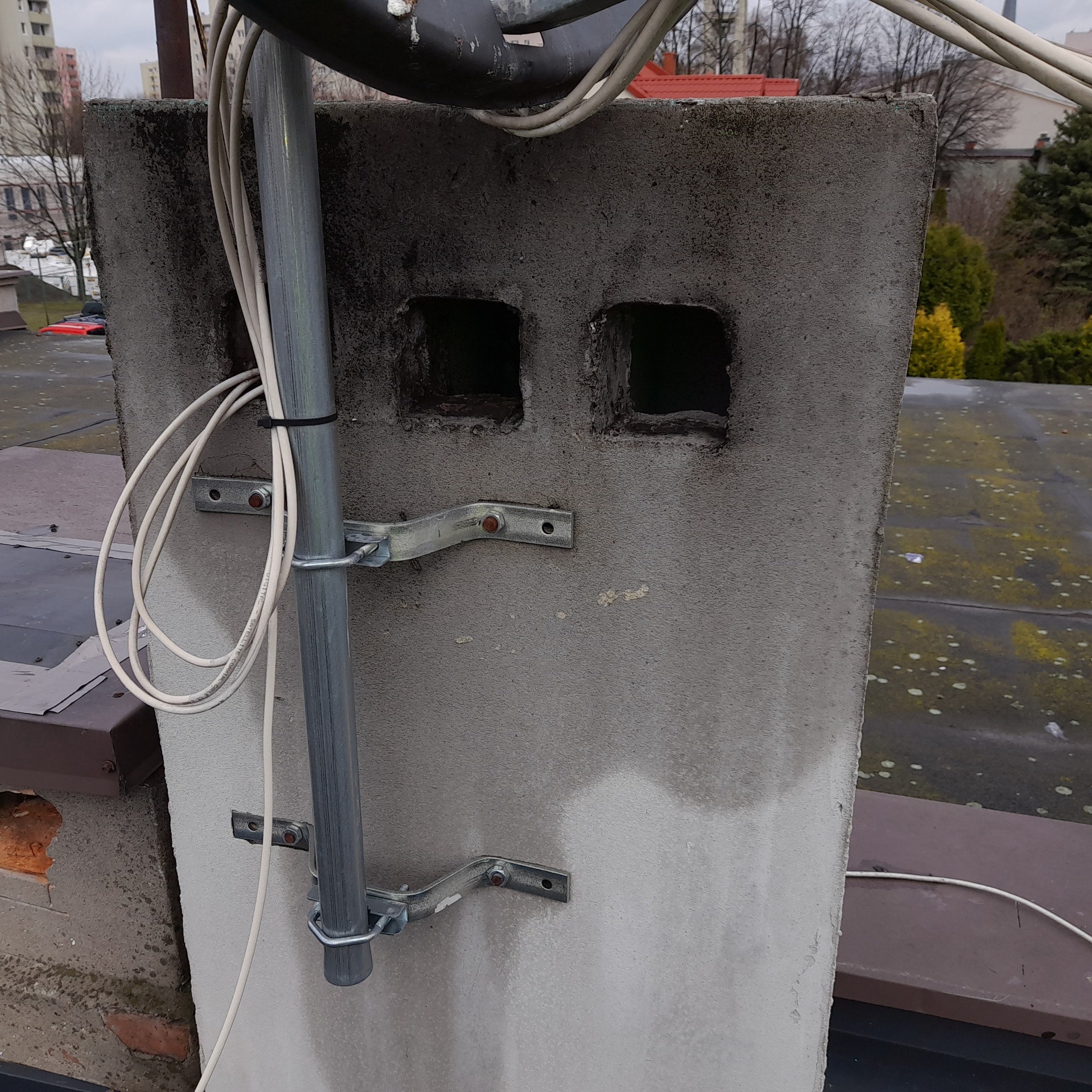 Szary komin wentylacyjny na dachu z widocznymi dwoma otworami, zamocowany metalowy maszt antenowy z przewodami. Dach pokryty plamami mchu.