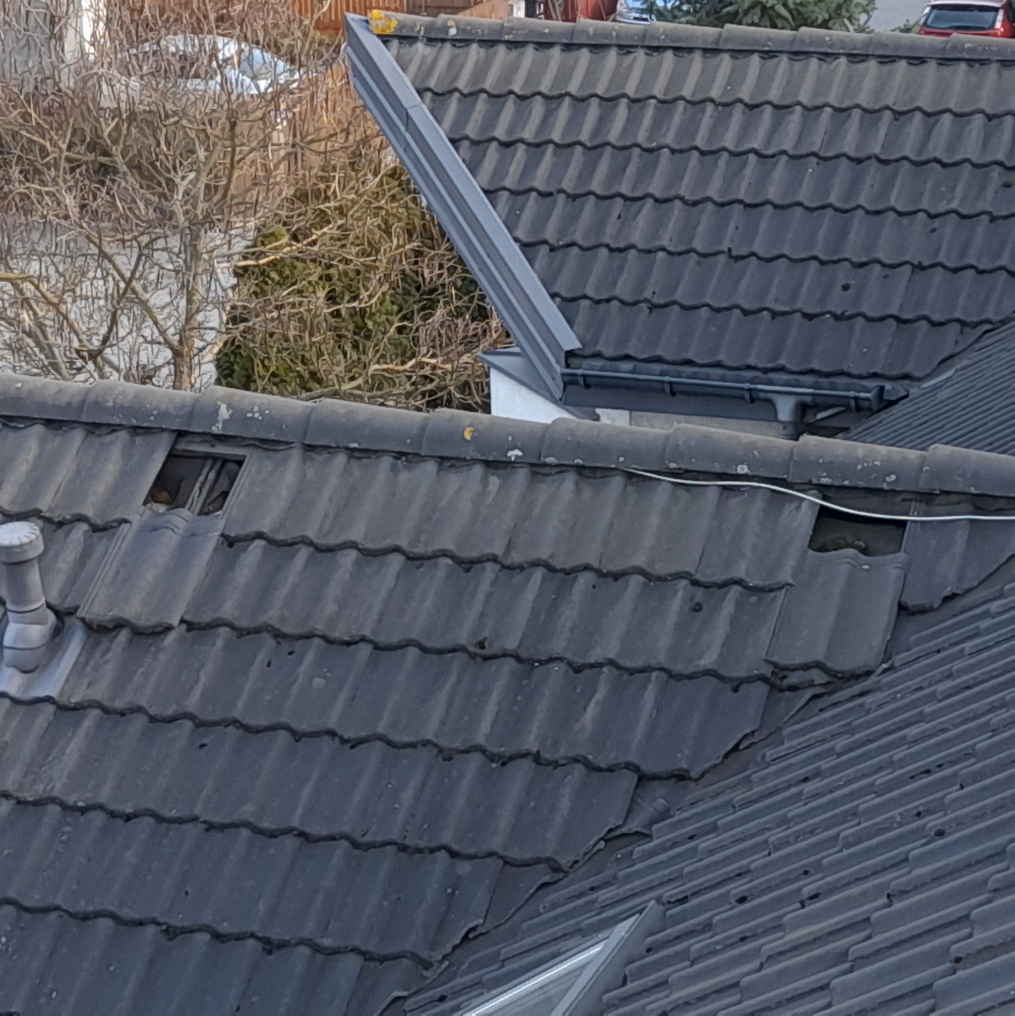 Uszkodzony dach z ciemnej dachówki ceramicznej, widoczna dziura i przesunięte płytki, w tle drzewo bez liści i fragment budynku z rynną.