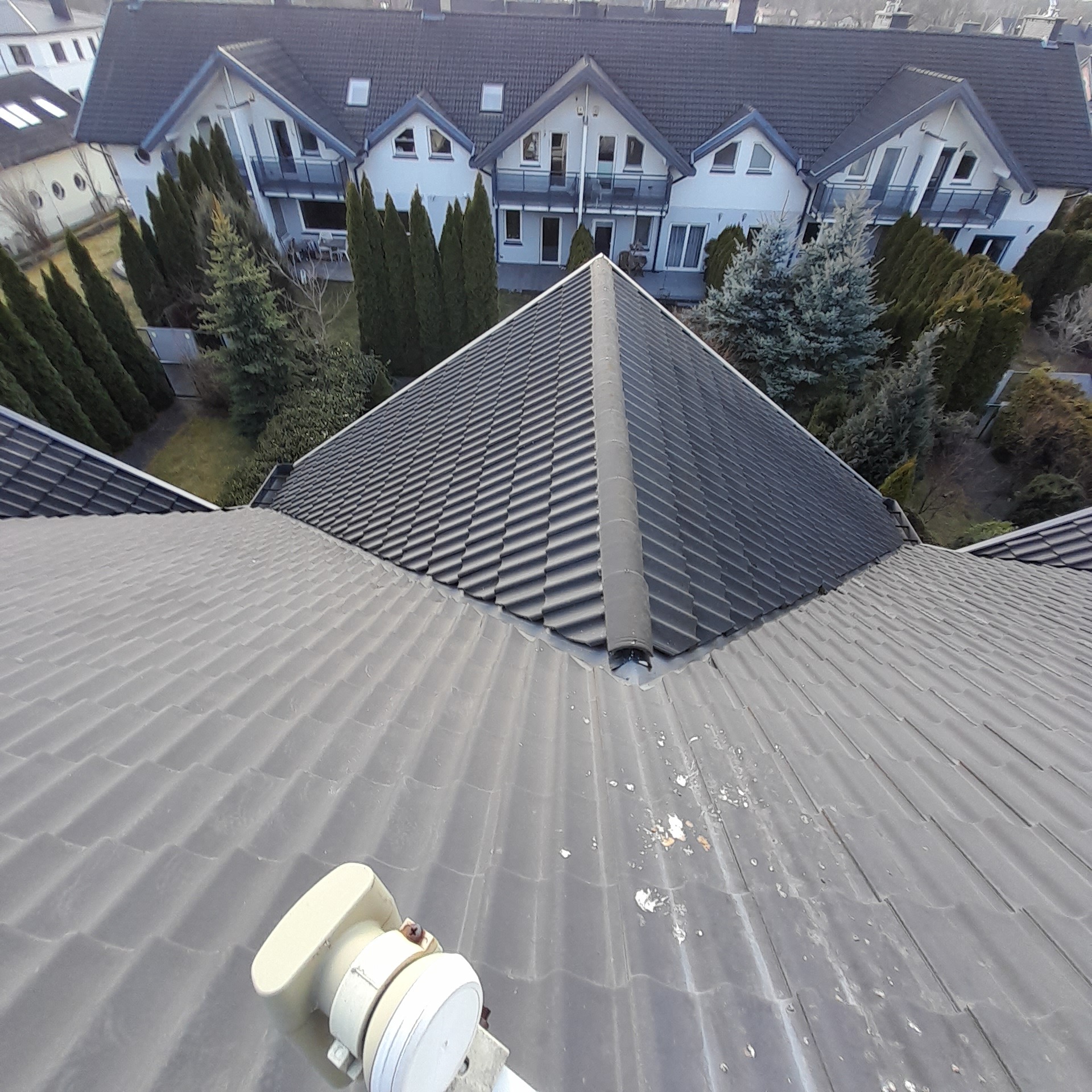 Widok z góry na szary dach z dachówki ceramicznej, z widocznym kominem i anteną satelitarną, w tle zabudowa jednorodzinna z zielenią.