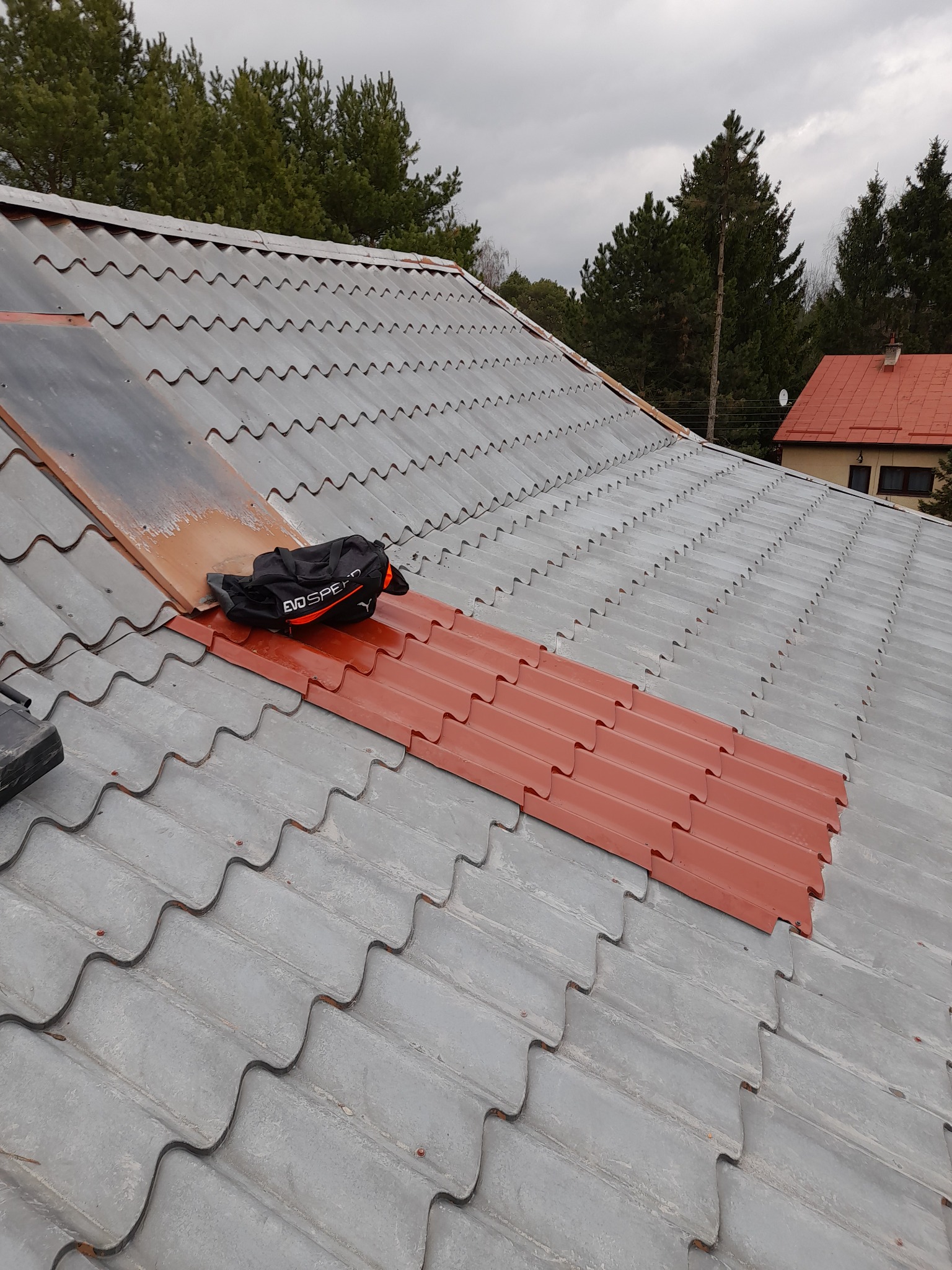 Fragment dachu z szarej blachodachówki, z widocznym obszarem świeżo pomalowanym na czerwono, na którym leży czarna torba sportowa EVO SPEED