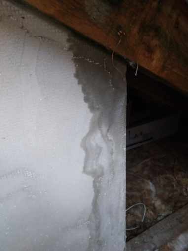 Biała ściana w poddaszu z widocznymi śladami przeciekania wody, brudne zacieki na styku z drewnianą belką stropową i izolacją.