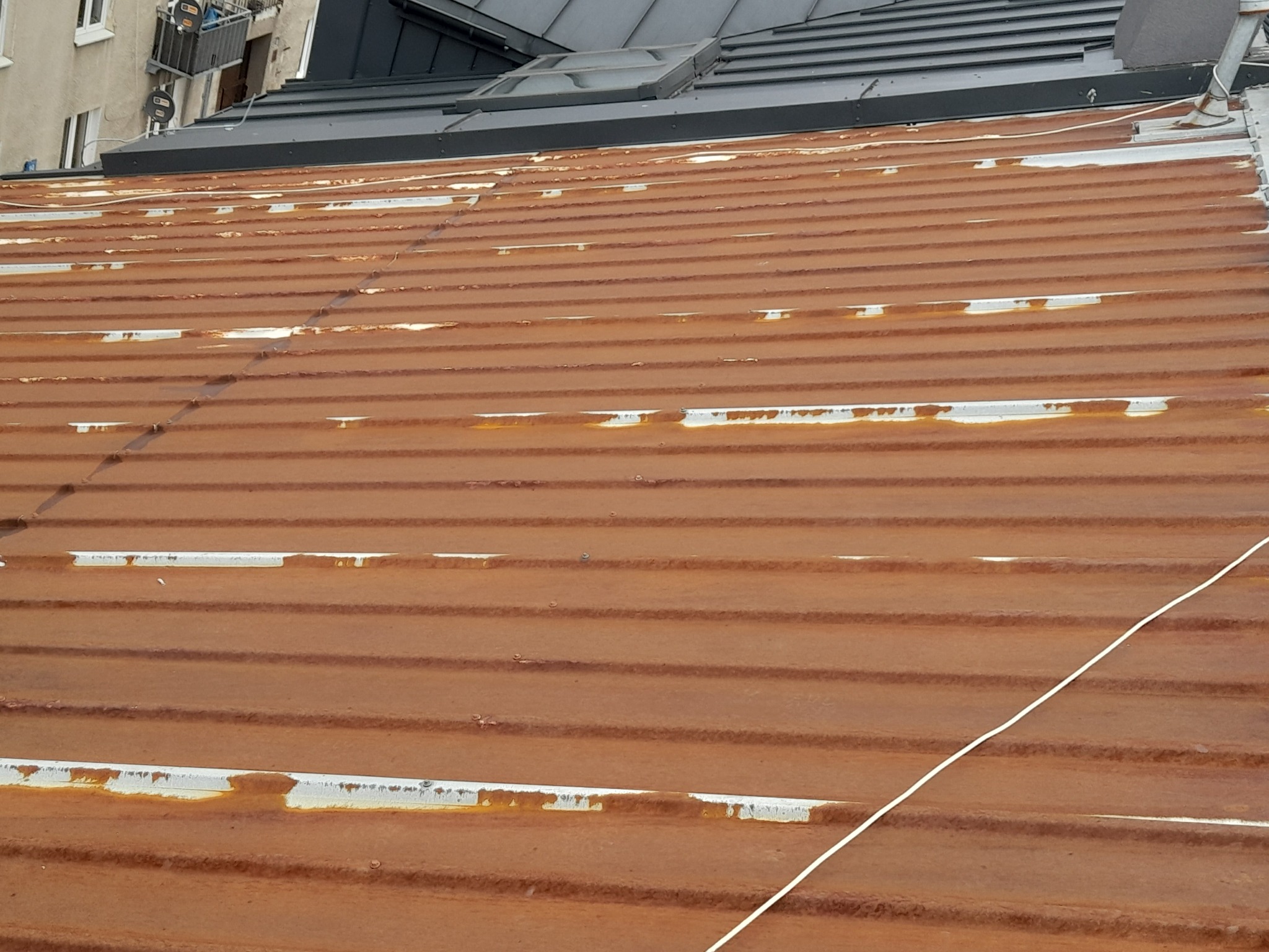 Zardzewiały blaszany dach z widocznymi śladami korozji i odchodzącą farbą, fragment budynku w tle.
