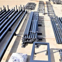 Elementy stalowe pomalowane na czarno, prawdopodobnie do budowy hali, leżące na betonowym placu w słoneczny dzień. Widoczne różne profile i kształtowniki, niektóre z przyspawanymi podstawami.