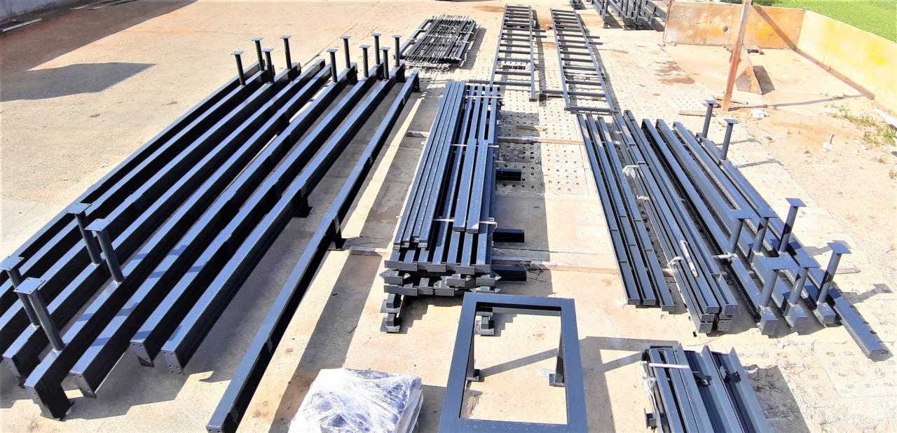 Elementy stalowe pomalowane na czarno, prawdopodobnie do budowy hali, leżące na betonowym placu w słoneczny dzień. Widoczne różne profile i kształtowniki, niektóre z przyspawanymi podstawami.