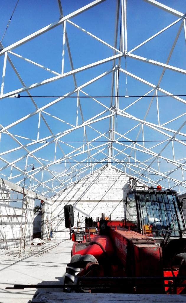 Wnętrze hali w trakcie budowy, widoczna biała stalowa konstrukcja dachu na tle błękitnego nieba oraz czerwony wózek widłowy z przodu.