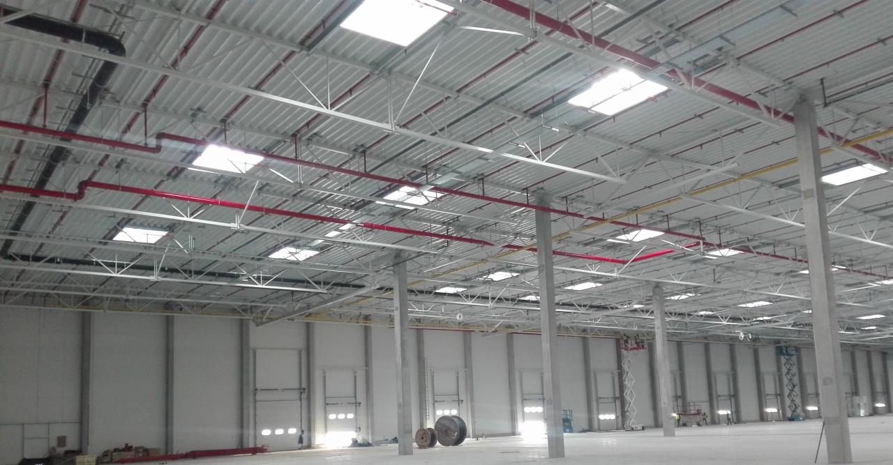 Wnętrze hali magazynowej w trakcie budowy, widoczne stalowe słupy, konstrukcja dachu z świetlikami, instalacje w kolorze czerwonym i żółtym, oraz zwinięte kable na podłodze.