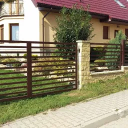 Brązowe, poziome przęsła ogrodzeniowe z prostokątnymi profilami, zamontowane na niskim, kamiennym murku z jasnego piaskowca, na tle domu z czerwonym dachem.