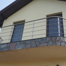 Szymon Zyskowski VESPER - Balkon z zaokrągloną, kamienną balustradą i prostą, metalową konstrukcją zabezpieczającą, widok z dołu na tle jasnego nieba.