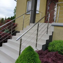 Schody wejściowe do domu z balustradą z polerowanej stali nierdzewnej, widok z dołu, z ozdobnymi krzewami i drzewami w tle.