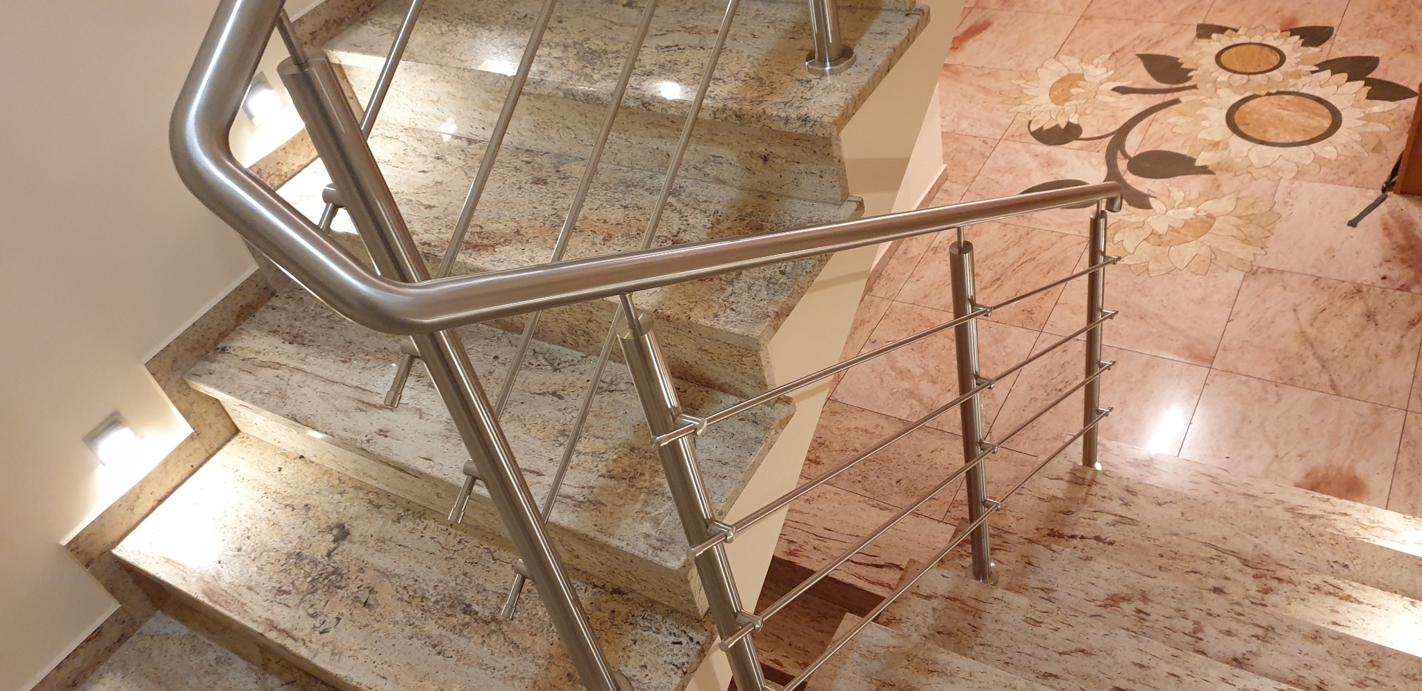 Schody wyłożone granitem z błyszczącą, metalową poręczą. Na podłodze widoczny wzór z płytek w kształcie kwiatów.