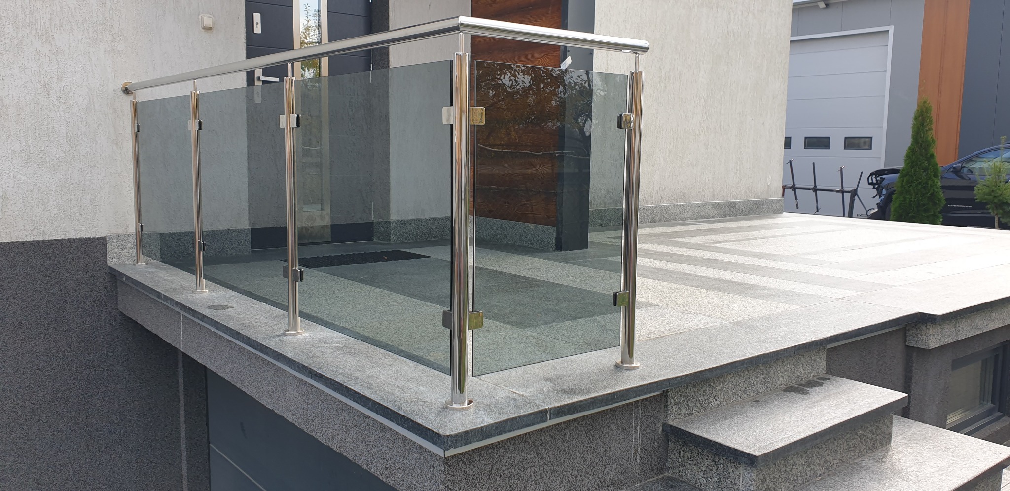 Szklana balustrada z elementami ze stali nierdzewnej na tarasie przed domem z wejściem po schodach, widoczne mocowania szyb.