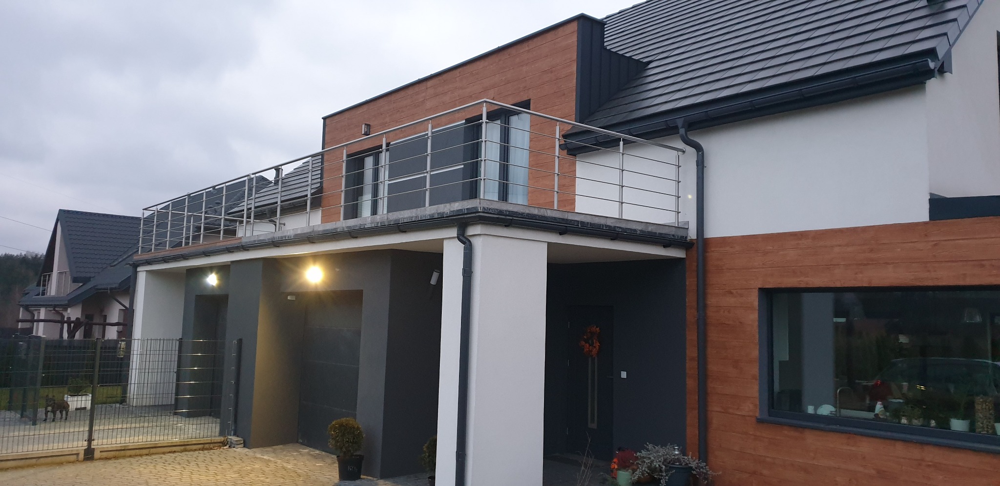 Nowoczesny dom z balkonem zabezpieczonym balustradą ze stali nierdzewnej, widok z zewnątrz, szara kostka brukowa na podjeździe.