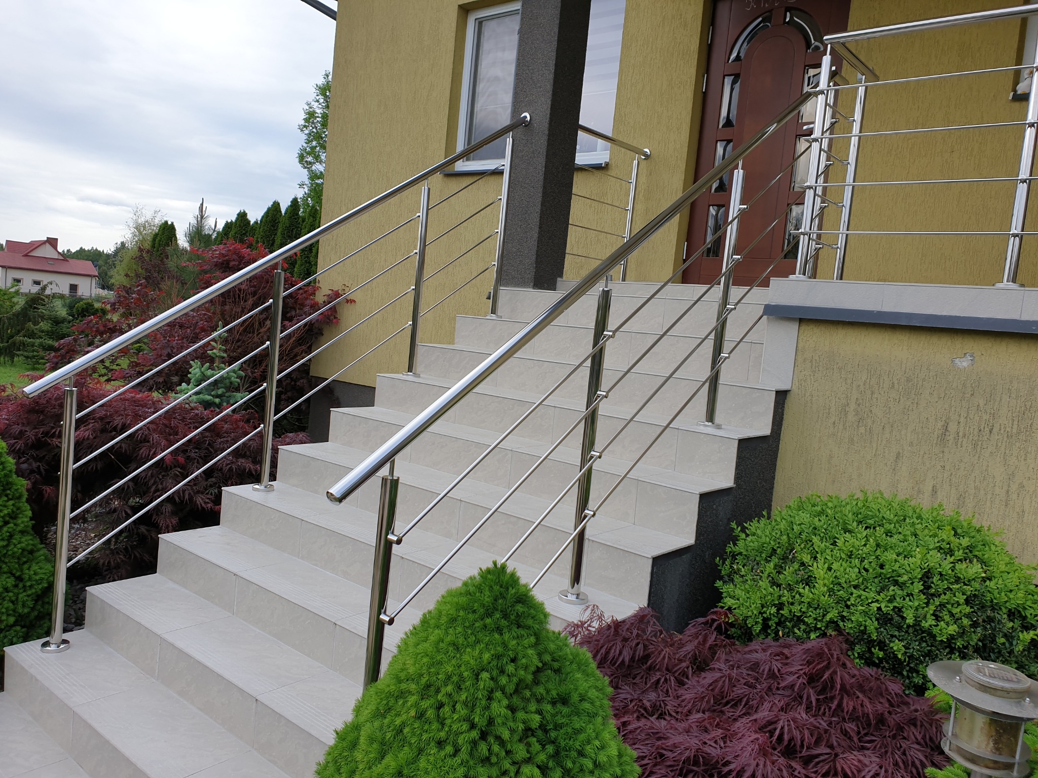 Schody wejściowe do domu z balustradą z polerowanej stali nierdzewnej, widok z dołu, z ozdobnymi krzewami i drzewami w tle.