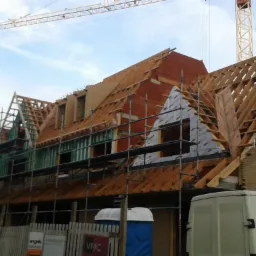 Budowa domu w trakcie: widoczne rusztowania, częściowo ukończona konstrukcja dachu z drewnianymi elementami i izolacją, na pierwszym planie samochód dostawczy i przenośna toaleta.