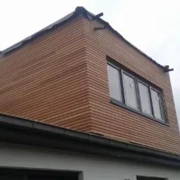 Fragment elewacji budynku z drewnianą okładziną w odcieniach brązu, widoczne okna z czarnymi ramami, rynna i fragment dachu sąsiedniego budynku na tle pochmurnego nieba.