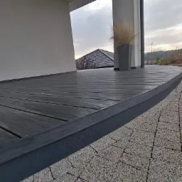 Nowoczesny taras z desek kompozytowych w odcieniach szarości, z zaokrąglonym brzegiem i donicą z trawą ozdobną, przylegający do jasnego budynku z widokiem na wiatrak w tle.