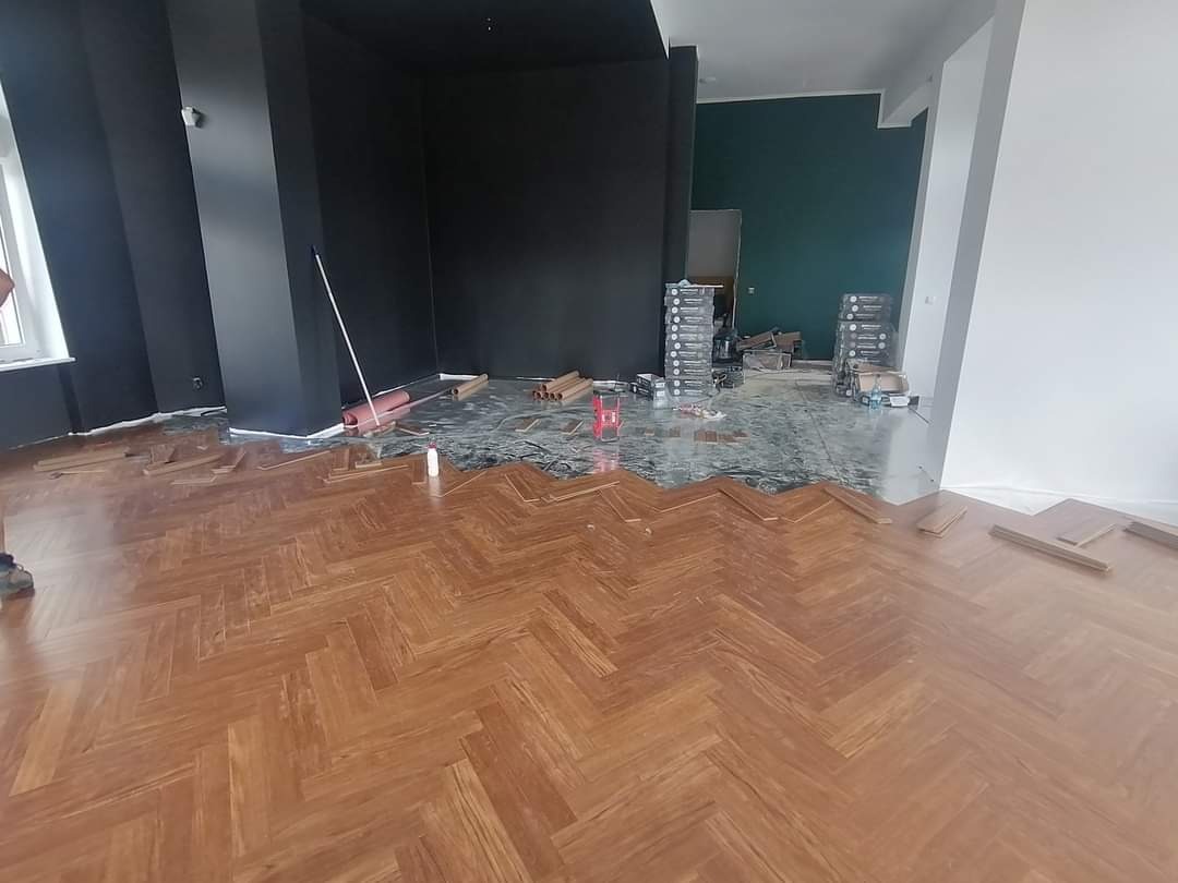 Układanie parkietu w jodełkę w pokoju z czarnymi i zielonymi ścianami, widoczne paczki paneli i narzędzia.