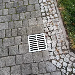 Szara kostka brukowa z metalową kratką odpływową w centrum, obok fragmentu chodnika z drobnej kostki granitowej i trawy.