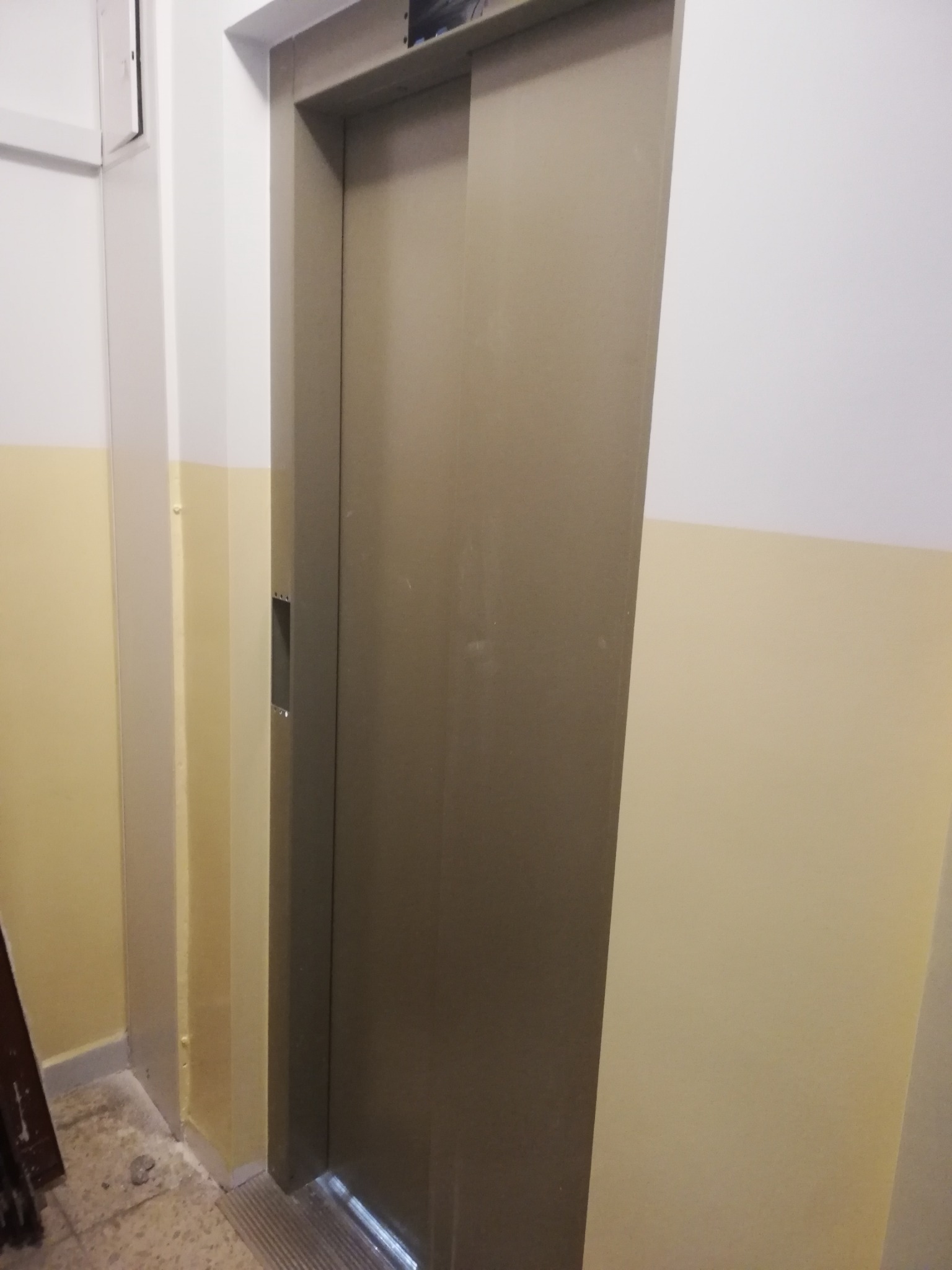 Zamknięte, metalowe drzwi windy w korytarzu z pomalowanymi na biało i żółto ścianami, widoczna kratka wentylacyjna pod drzwiami.