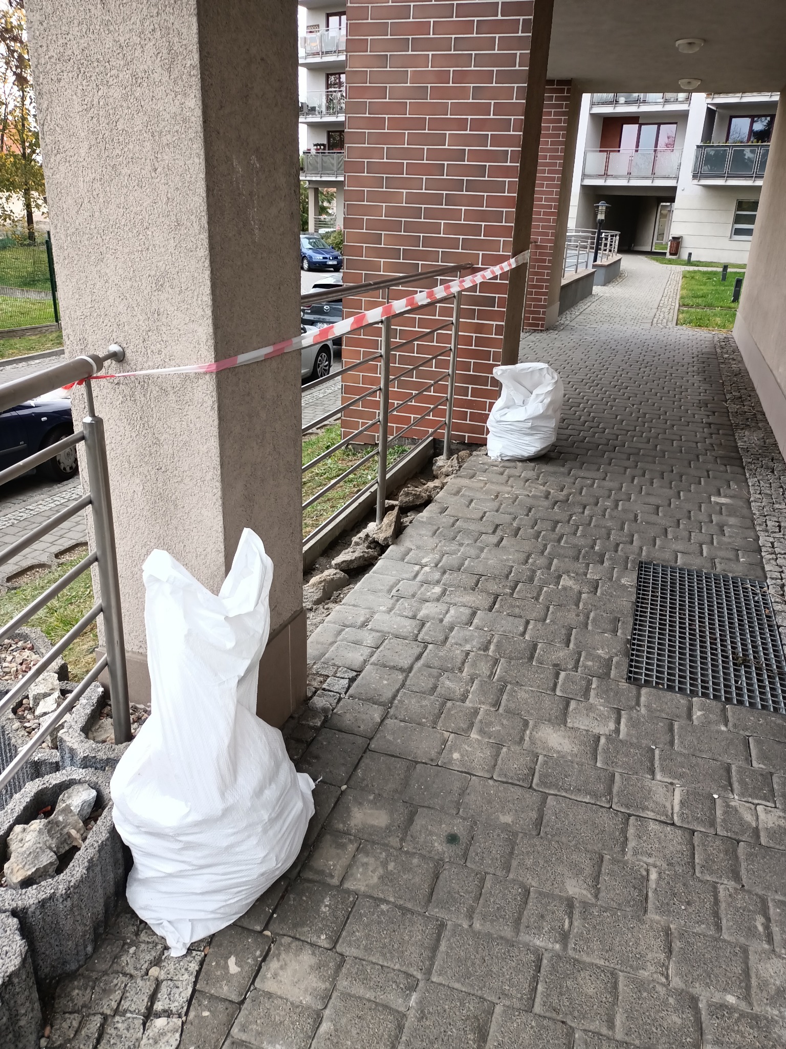 Fragment chodnika z kostki brukowej w Gdańsku, zabezpieczony taśmą, z workami gruzu i elementami obrzeża w trakcie prac.