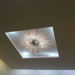 Sufit podwieszany z kwadratową wnęką oświetloną LED-ami, w której umieszczony jest nowoczesny żyrandol z elementami przypominającymi papryczki chili, rzucający cienie na teksturowaną powierzchnię.