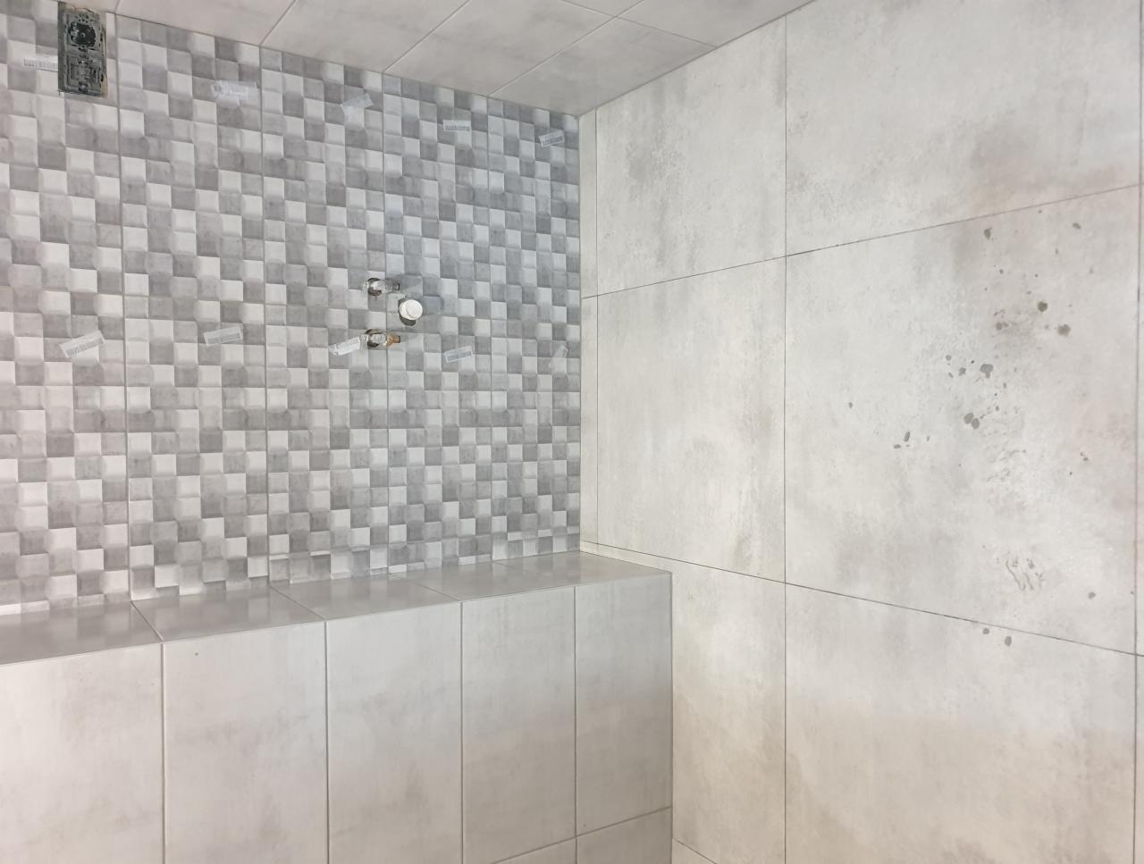 Ściana łazienki wyłożona szarymi płytkami w dwóch odcieniach, z widocznymi przyłączami do armatury i pozostałościami po naklejkach zabezpieczających, sąsiadująca z gładką, jasnoszarą ścianą...