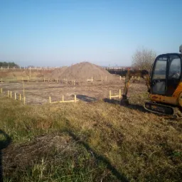 Żółta mini koparka stoi na działce z wyznaczonym obrysem fundamentów pod budynek, w tle pryzma ziemi i budynek w trakcie budowy.